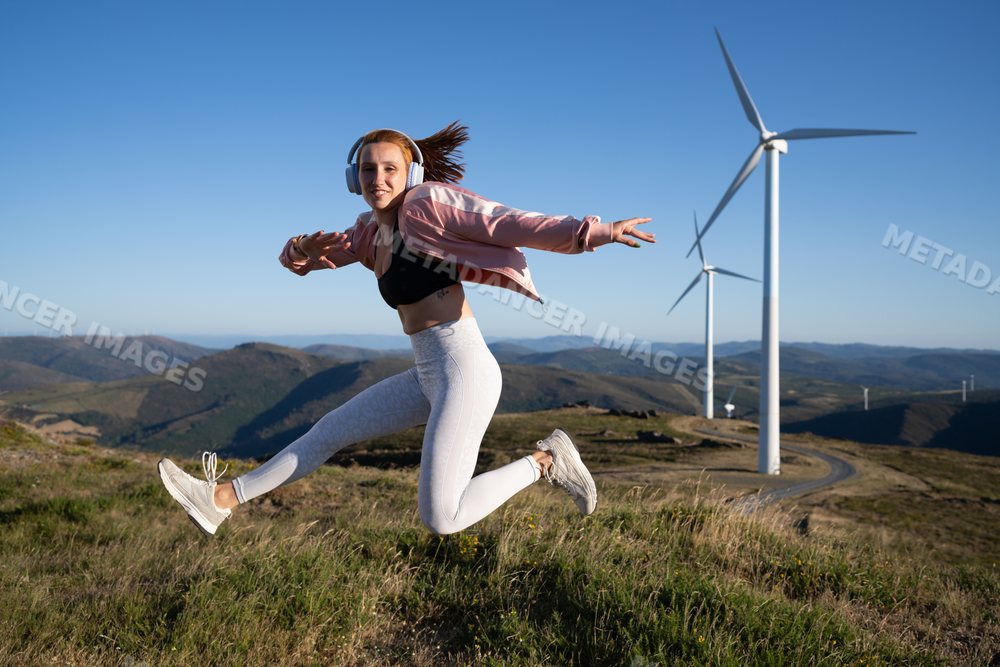 A young white woman jumps energetically outdoors near wind turbines