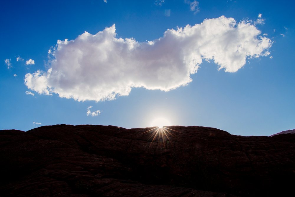 Sunset with sun partially visible over silhouetted rock formations and clouds in the blue sky at red Cliffs Desert State Park, Snow Canyon, Utah, USA.
