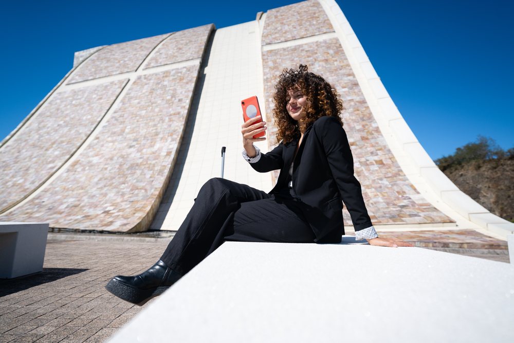 businesswoman sitting on a bench taking a selfie outside a modern architectural building during a business trip, Spain, Santiago De Compostela.