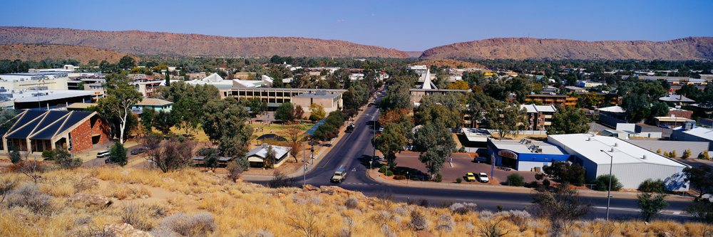 Panoramic townscape view of Alice Springs with buildings, roads, trees, dry terrain, and mountain ranges under a clear blue sky, Alice Springs Northern Territory, Australia.