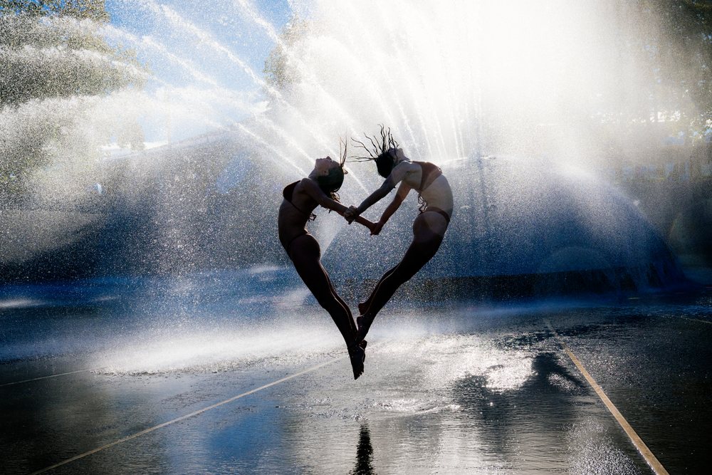 Two women jumping and holding hands in a dynamic acrobatic pose in front of a water fountain spray, partner, acrobatics, yoga, USA.