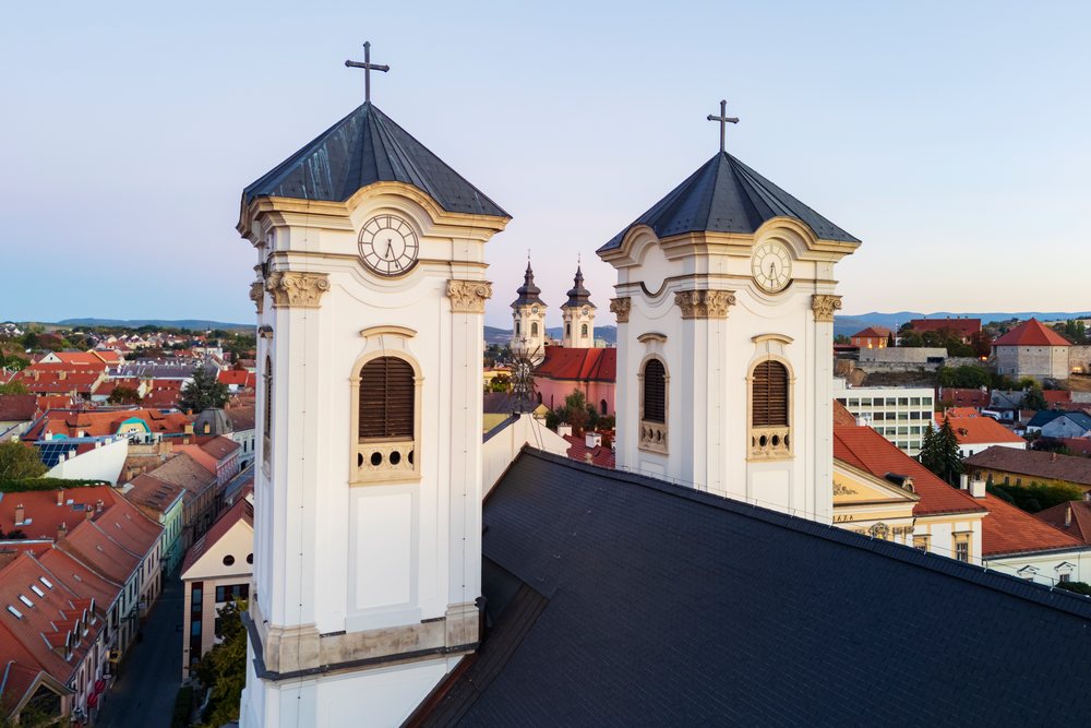 The Minorite Church towers frame the distant twin towers of Egri Barátok Temploma within a cityscape of Eger, at sunset under a clear sky, Hungary.