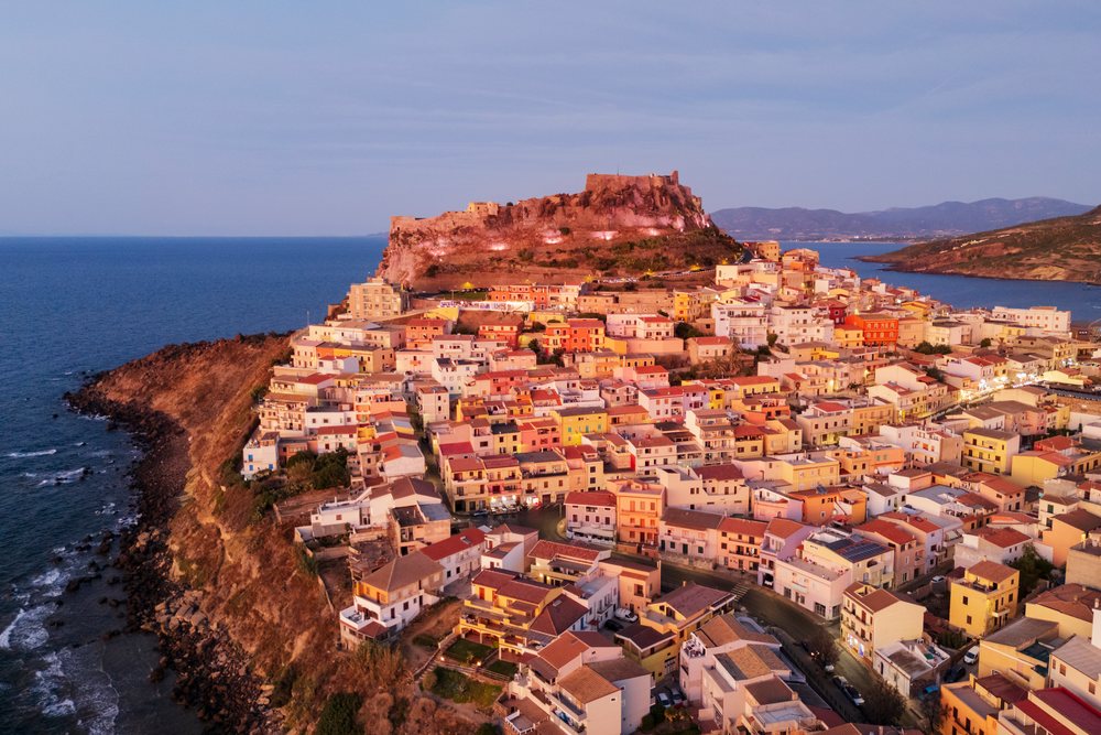 Hilltop Citadel and dense village cluster on the rocky coast of Castelsardo at dusk with illuminated buildings and Mediterranean Sea backdrop, Travel Images, Sardinia, Italy.