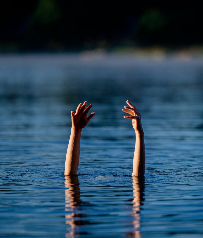 Two uplifted arms and hands emerging from the calm blue water surface of a lake at sunset, USA.