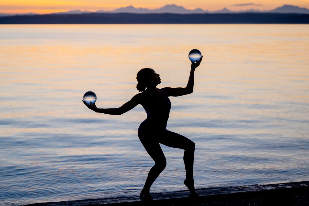 Silhouette of a woman balancing in a yoga acrobatic pose while holding two spherical objects by the water at sunset, USA.