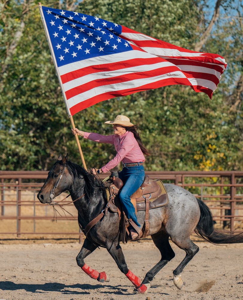 Woman rides a horse outdoors holding a large American flag in the Southwest USA setting.