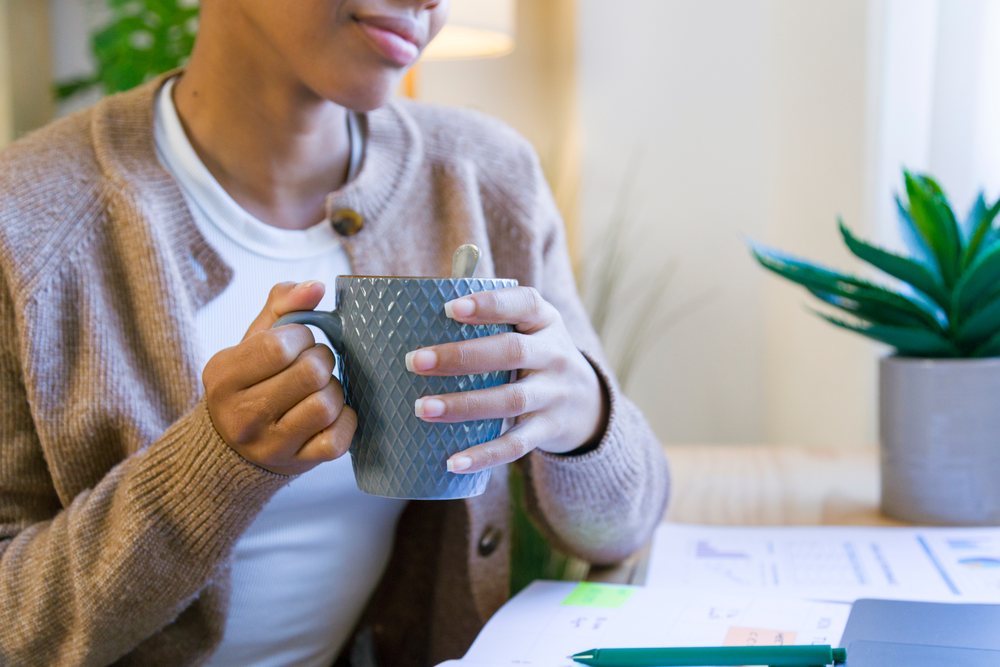 Woman holding a gray patterned cup with a spoon inside at her home office desk with paperwork, pen, and potted indoor plant nearby, remote work.