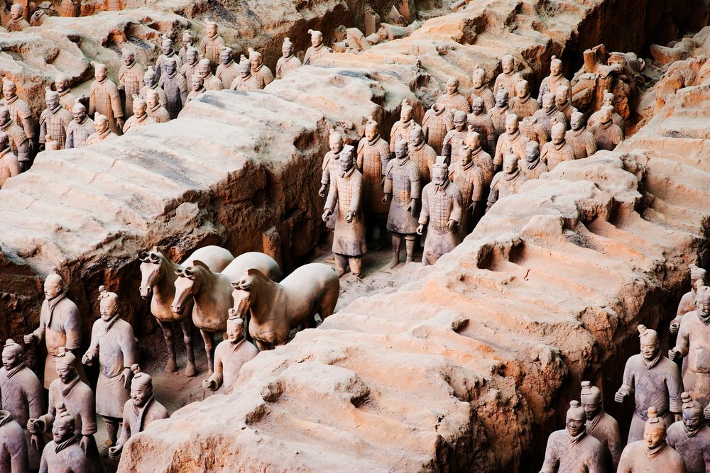 Life-sized terracotta warriors and horses with detailed armor and hairstyles are arranged in excavation trenches at an indoor archaeological site, Shaanxi Province, China.