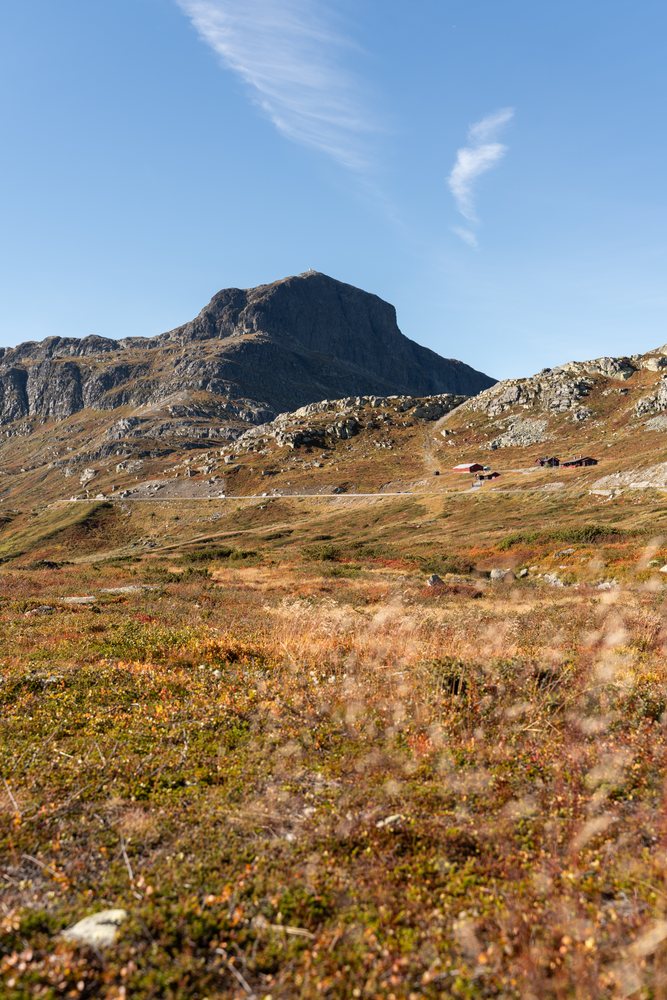 Mountain landscape featuring rocky terrain, hillside vegetation, and rural houses under a clear blue sky, Norway.