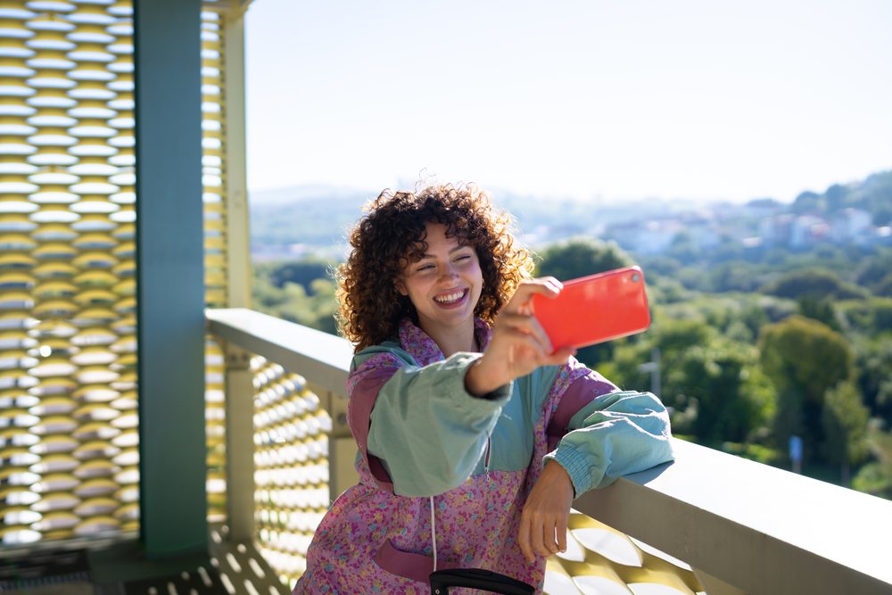 Woman with curly hair taking a selfie on an outdoor balcony in Spain during daylight, with urban scenery in the background.