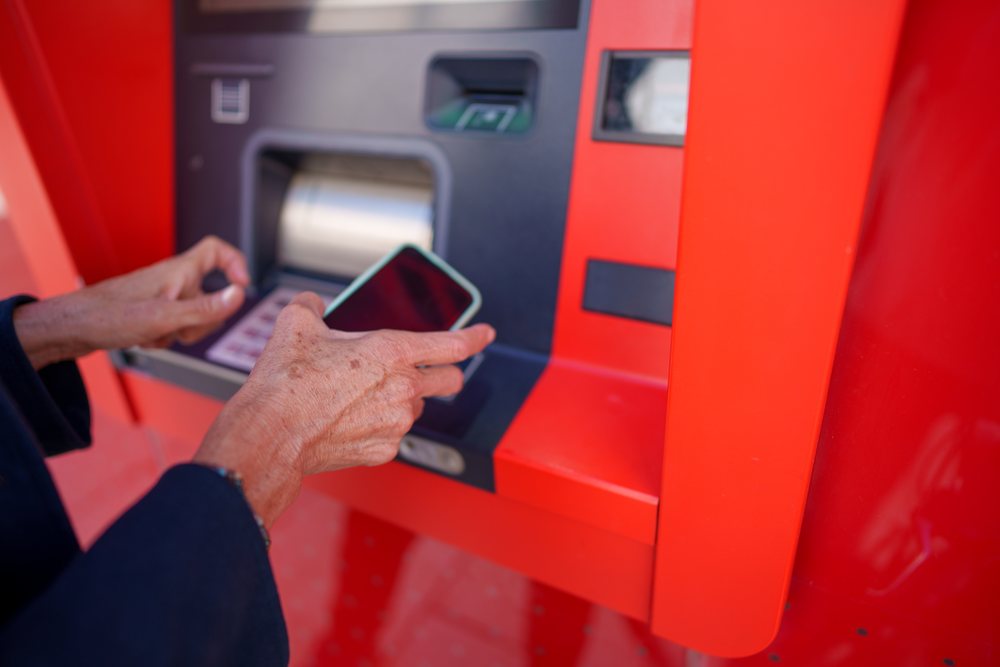 Hands holding a smartphone at a red ATM machine in Málaga performing a banking transaction outdoors.