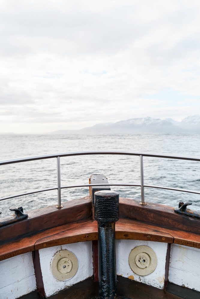 View from the bow of a wooden boat looking out over the ocean with distant mountains and a cloudy sky, Iceland.