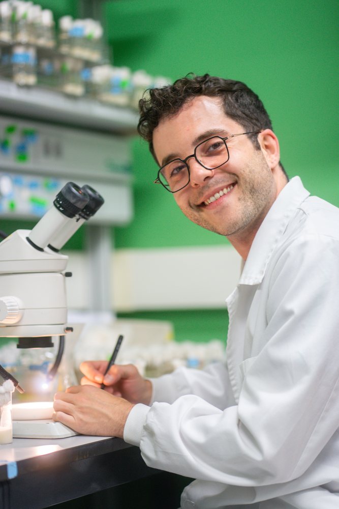 A male scientist wearing a lab coat is writing notes next to a microscope, science, biology, research, Inside A Research Laboratory.