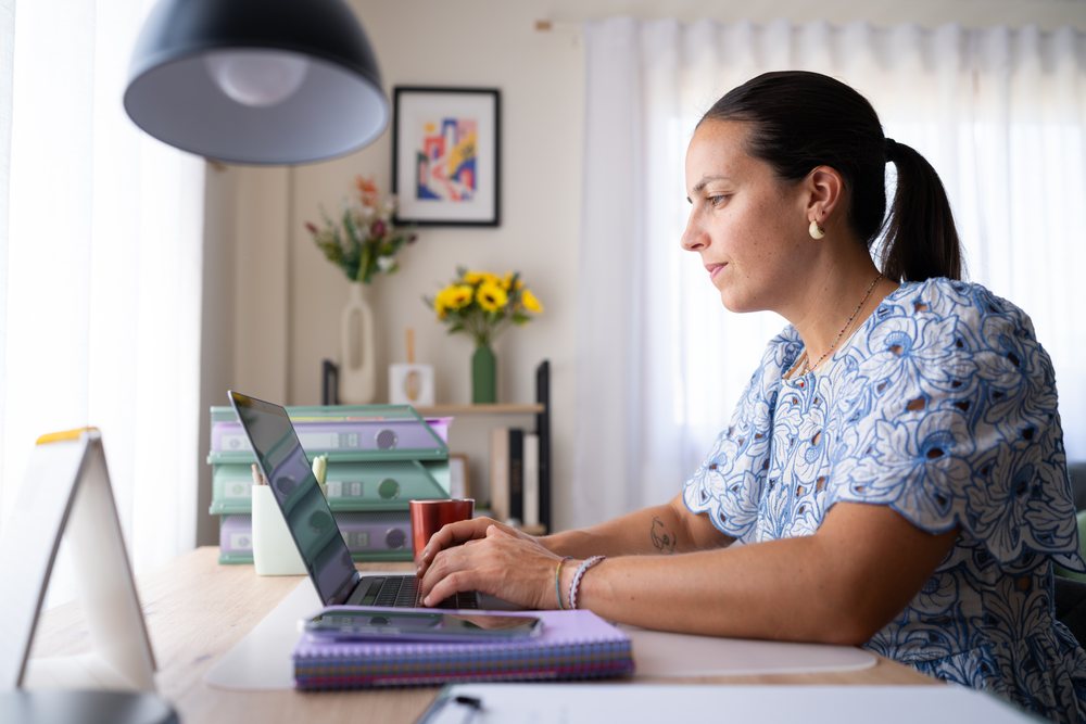 Woman typing on a laptop at an organized desk in a home office with natural light from a window, telecommuting, Spain.