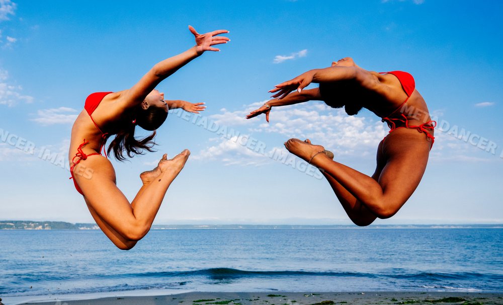 Two women jump in synchrony doing partner acrobatics on a beach