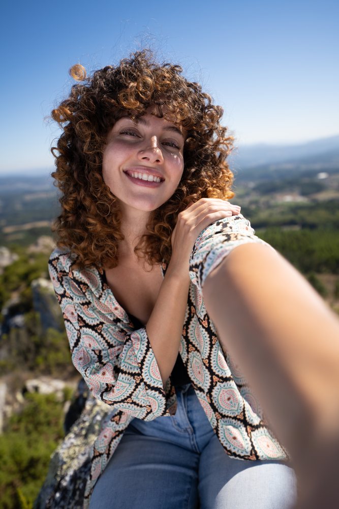 Young woman with curly hair wearing a patterned blouse and jeans takes a smiling selfie outdoors, with a scenic mountainous landscape, Pico Sacro, Galicia, Spain.