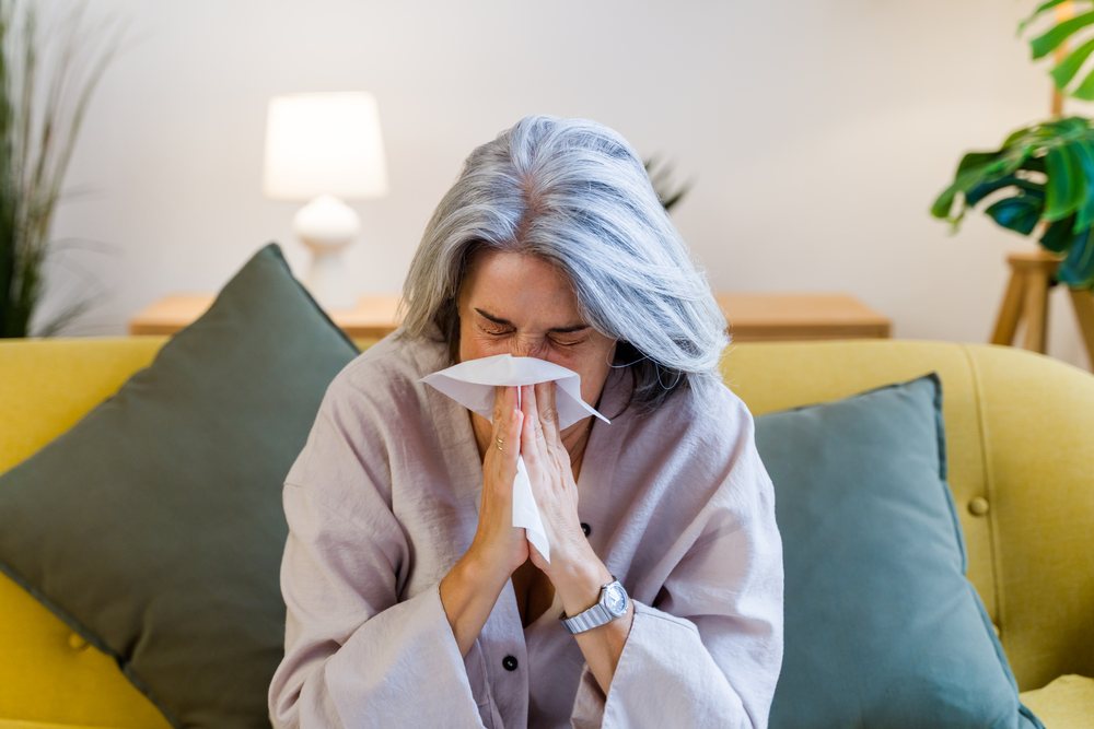 Woman with gray hair sneezing into a white tissue while sitting on a yellow couch with green cushions in a living room, Spain.