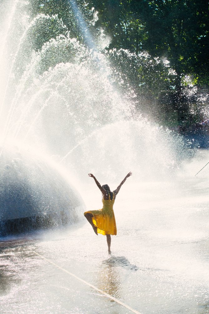Woman dancing joyfully in the water spray of a large outdoor fountain on a sunny day, USA.