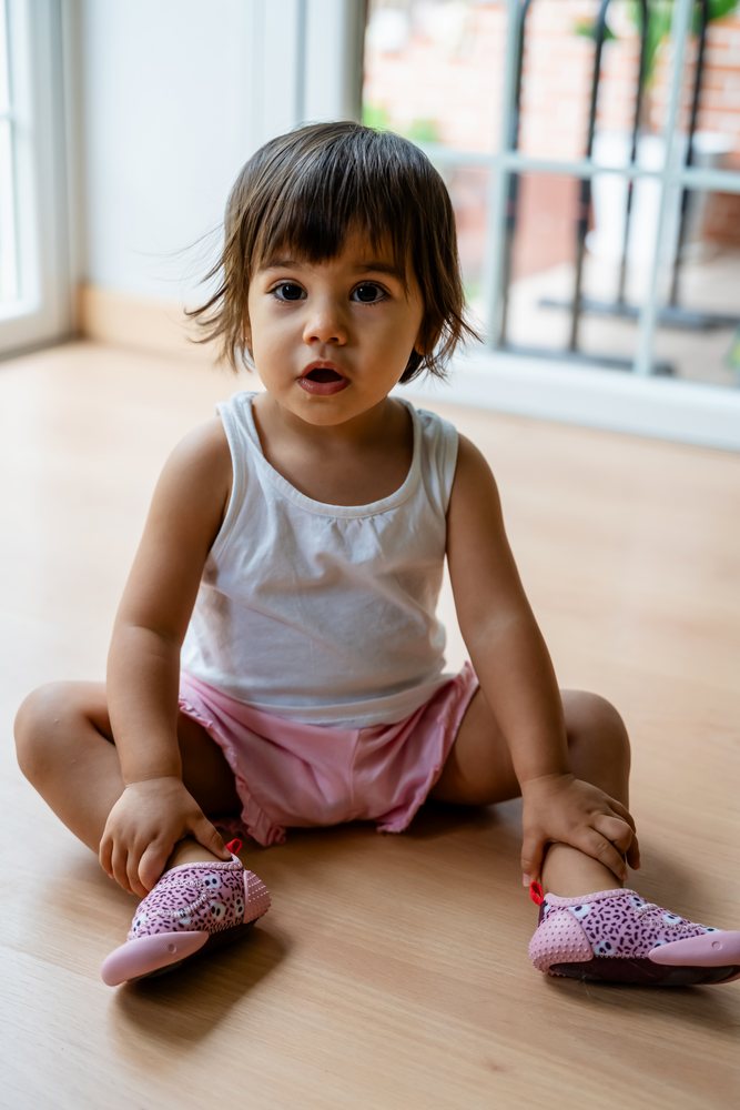 Toddler sitting on a wooden floor near a window indoors, wearing pink shoes and a white tank top, kids, Spain.