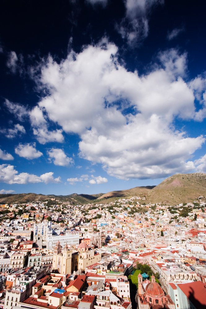 Colorful buildings of Guanajuato cityscape with mountains and cloudy blue sky in the background.