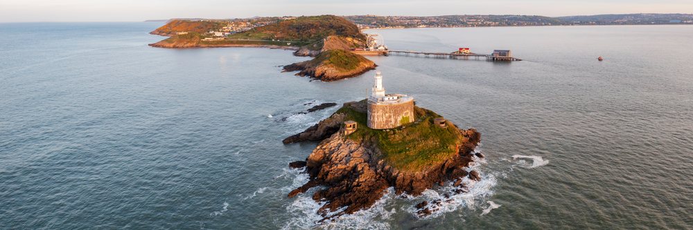 Mumbles Lighthouse stands on a rocky island at sunrise with a pier, ferris wheel, and coastline, Landscape, Swansea, Glamorgan, Wales.