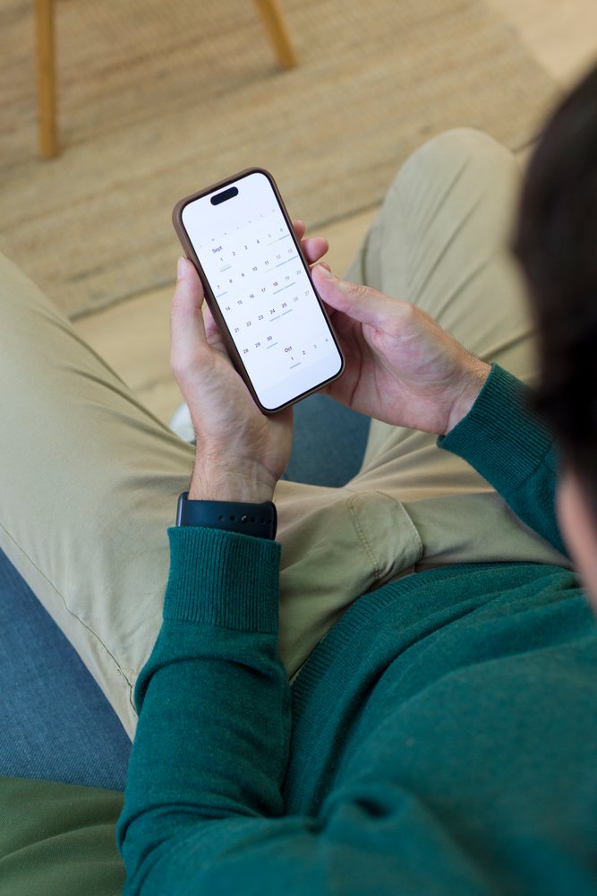 Person seated indoors viewing a calendar app on a smartphone held in hands, Young adult, home.