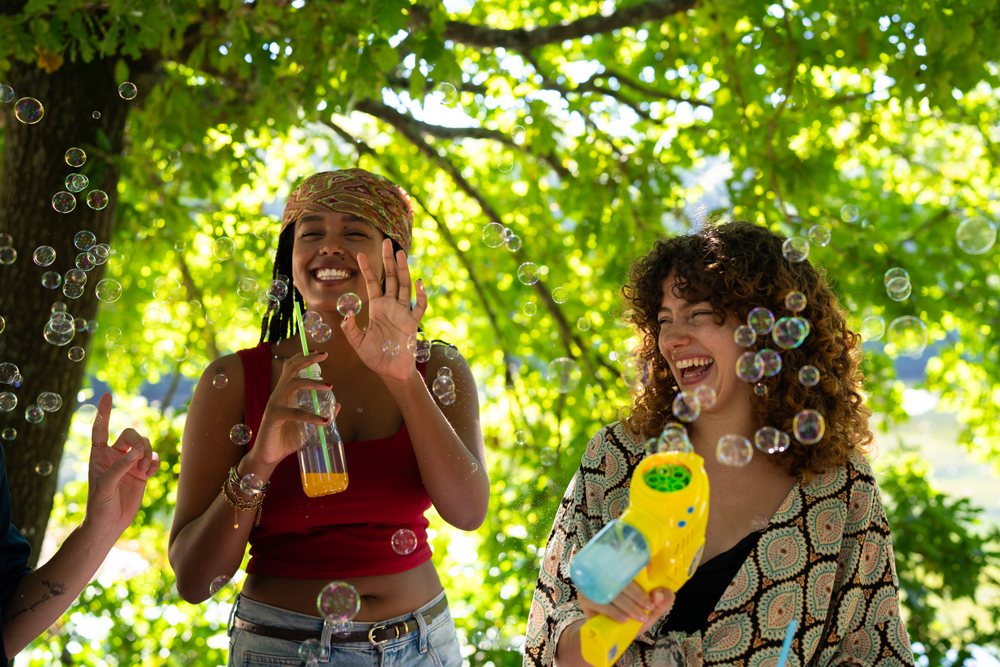 Two young women laugh together outdoors under a leafy tree playing with bubbles and holding drinks, Spain.