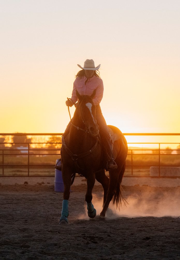 Rider wearing a cowboy hat rides a horse in a dusty outdoor arena at sunset, Southwest USA.