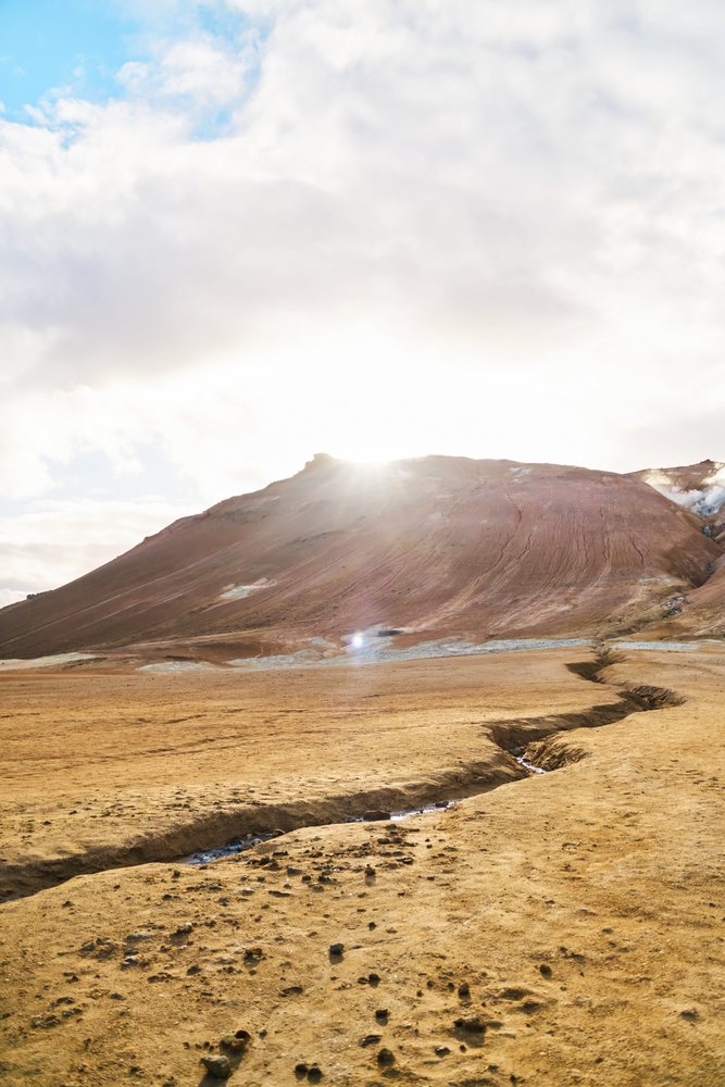 Sunlight shines over a volcanic landscape with cracked dry ground and geothermal activity, Iceland.