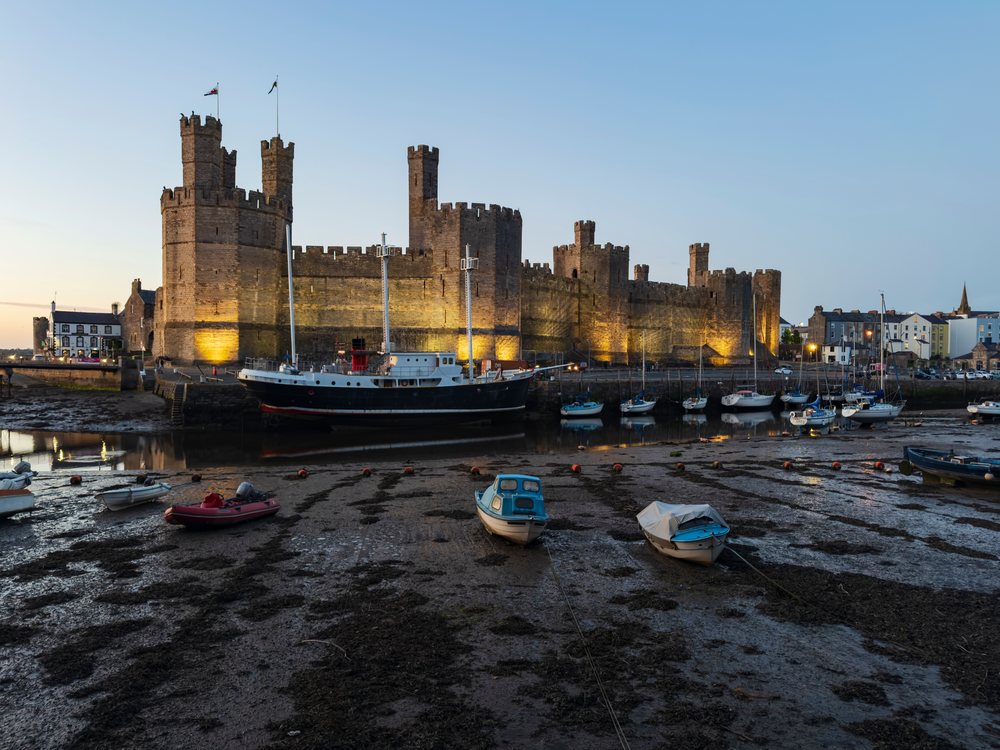 Caernarfon Castle illuminated at evening with boats moored in the harbor at low tide, Landscape, UK, Wales.