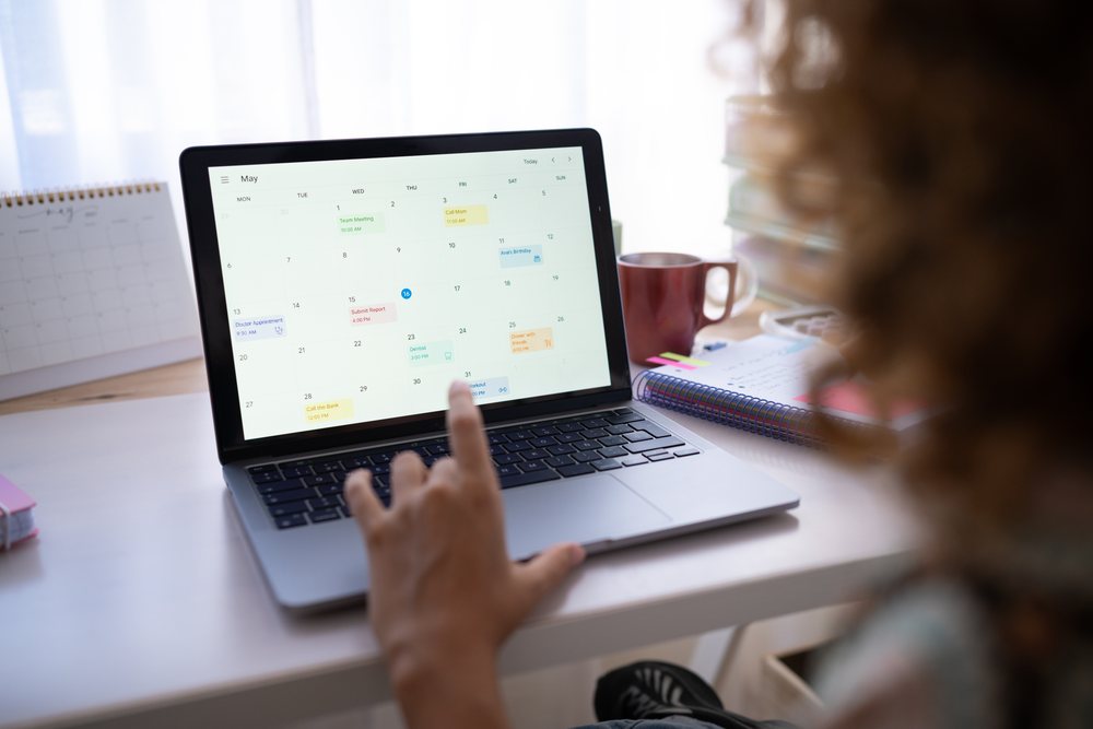 Woman using a digital calendar on a laptop at a desk with notebook and coffee mug in a home office, Spain.