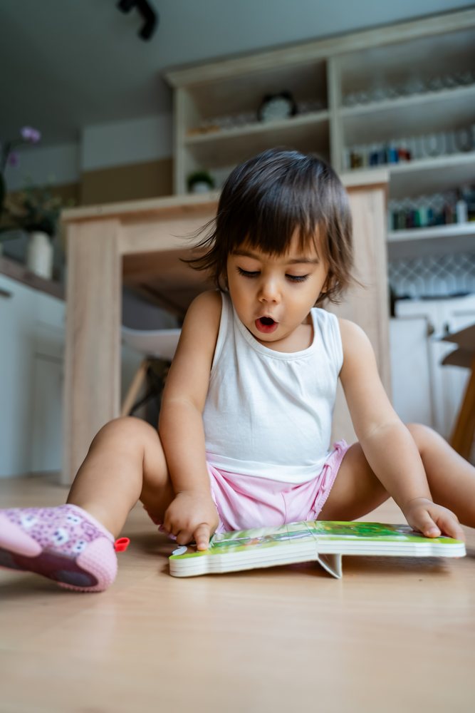 Toddler girl sitting on a wooden floor indoors at home, reading a colorful book, kids, Spain.