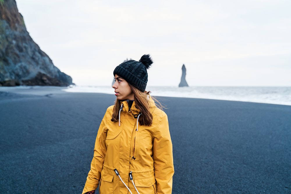A white woman in winter clothing stands on a black sand beach