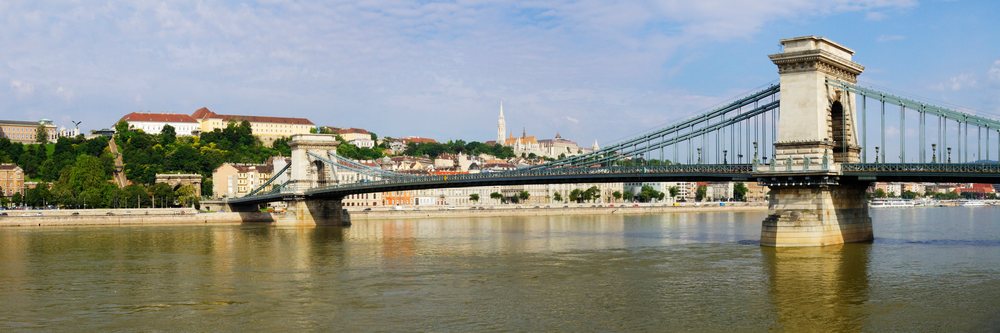 Chain Bridge spans the River Danube with urban buildings and a clear sky, reflecting in the water, Budapest, Hungary.