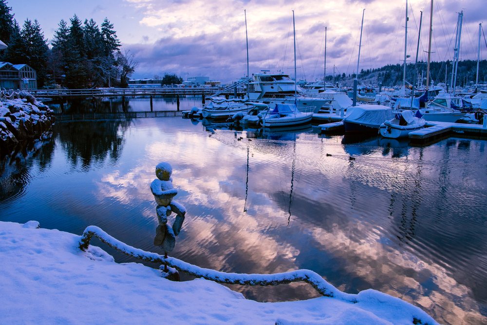 Boats docked at a marina, with snow covering the ground and a cloudy sky reflected in the calm water during a winter evening, WA, USA.
