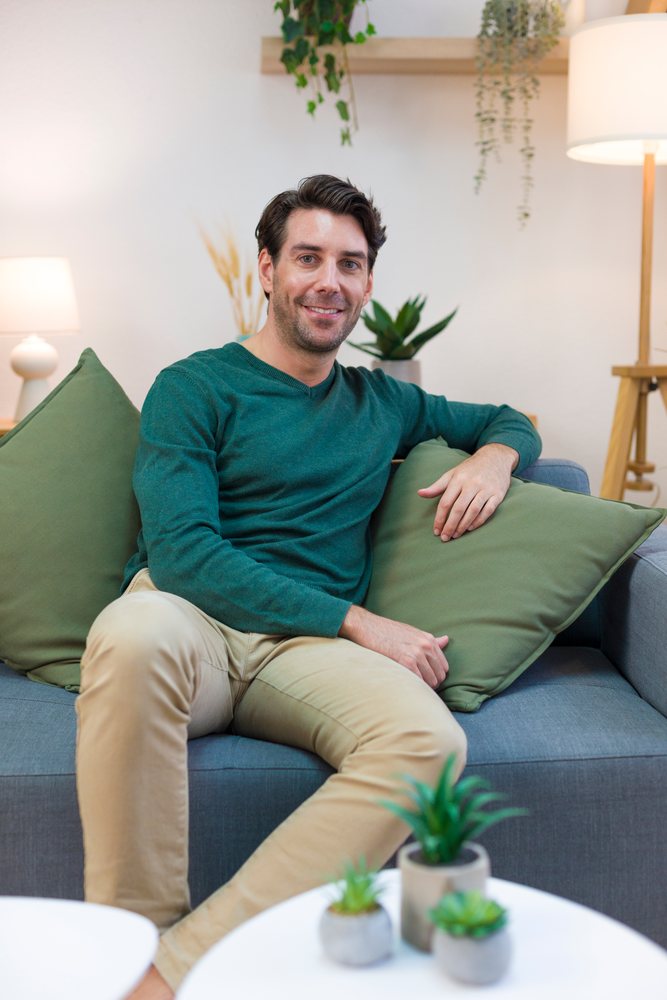 Man sitting on sofa holding a green pillow in a modern living room with indoor plants and table lamps, technology, young adult, home.