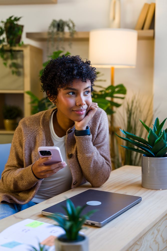 Woman with curly hair wearing a brown cardigan and white shirt holds a smartphone thoughtfully while seated at a wooden desk with a closed laptop and potted plants indoors, Spain.