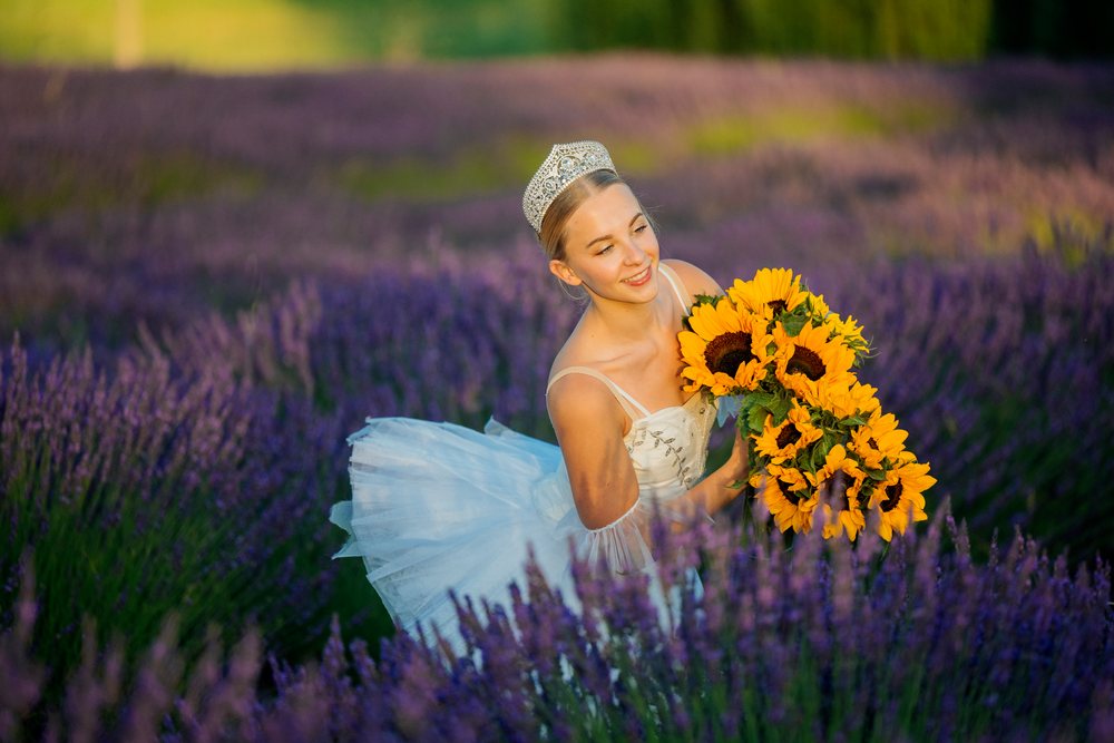 A ballet dancer wearing a silver tiara holds a bouquet of sunflowers while posing in a lavender field, WA, USA.