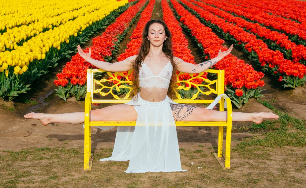 Tattooed woman wearing a white skirt and crop top performs splits while sitting on a yellow bench in a sunny tulip field, USA.