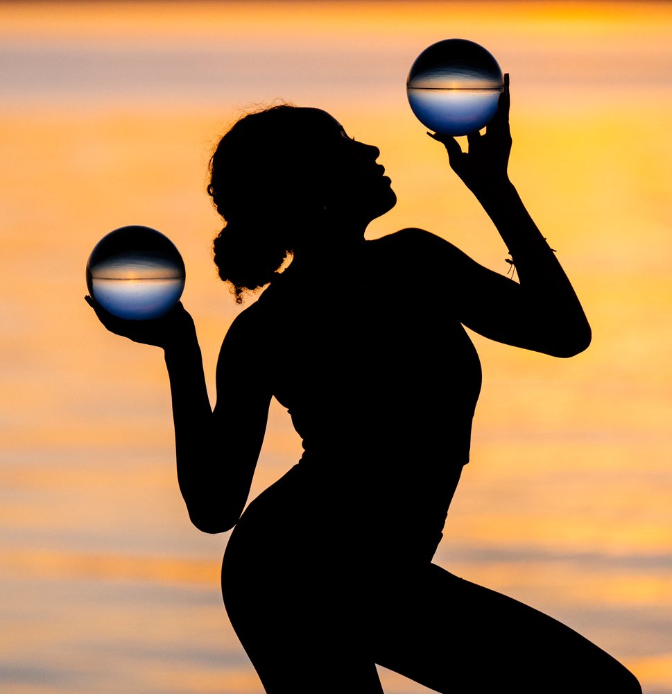 Silhouette of a woman holding two glass balls in a yoga-inspired pose with a sunset sky in the background, USA.