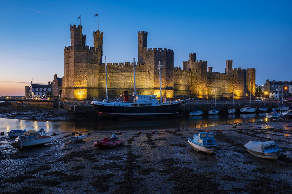 Stone castle walls with battlements and towers stand by the harbour at Low Tide at Dusk, Landscape, Caernarfon, Gwynedd, Wales.