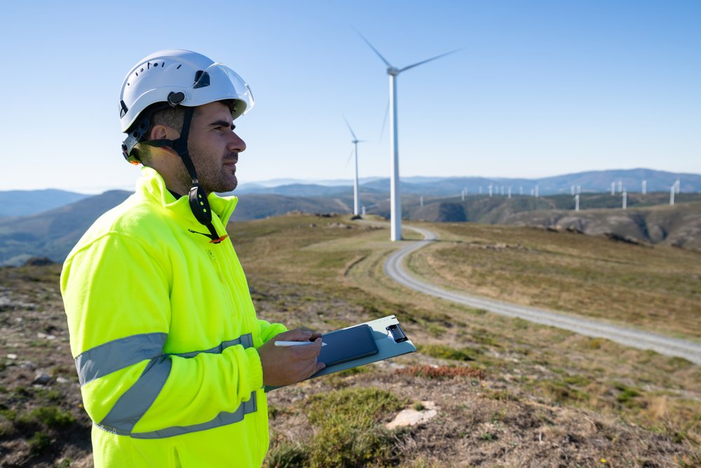 Engineer wearing a safety helmet and high visibility jacket inspects wind turbines on a grassy, hilly wind farm on a clear day, sustainability, renewable energy, Galicia, Spain.