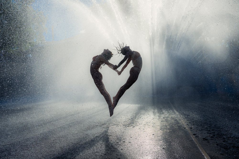 Two women jump and hold hands to form a heart shape while playing in water spray on a street, partner, acrobatics, yoga, USA.
