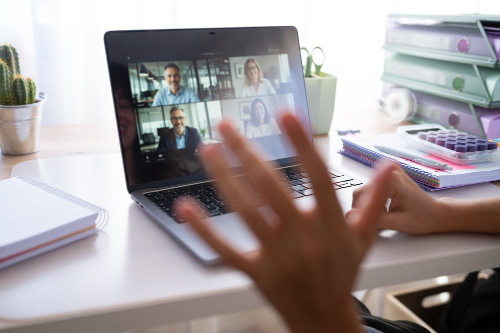 Hand waving during a video call on laptop at home office desk with notebooks, plant, and office supplies, Spain.