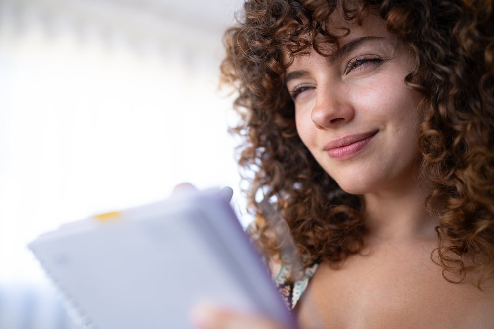 Woman with curly hair smiling and holding a spiral notebook indoors in bright natural light, home, girl, Spain.
