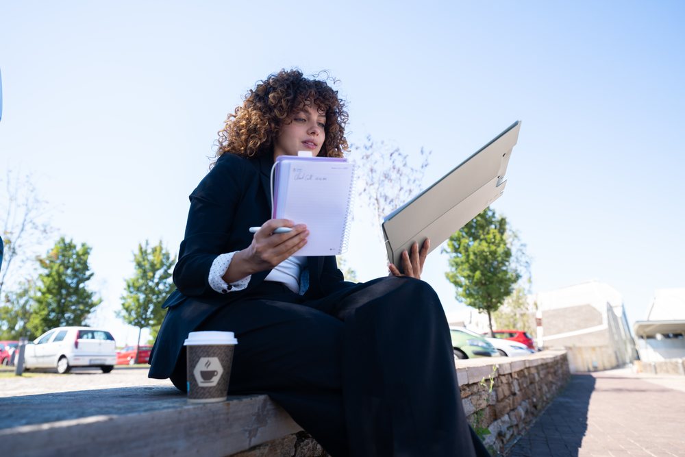 Woman writing notes and holding a laptop while sitting outdoors in an urban setting with a coffee cup nearby, businesswoman, traveling executive, Spain.