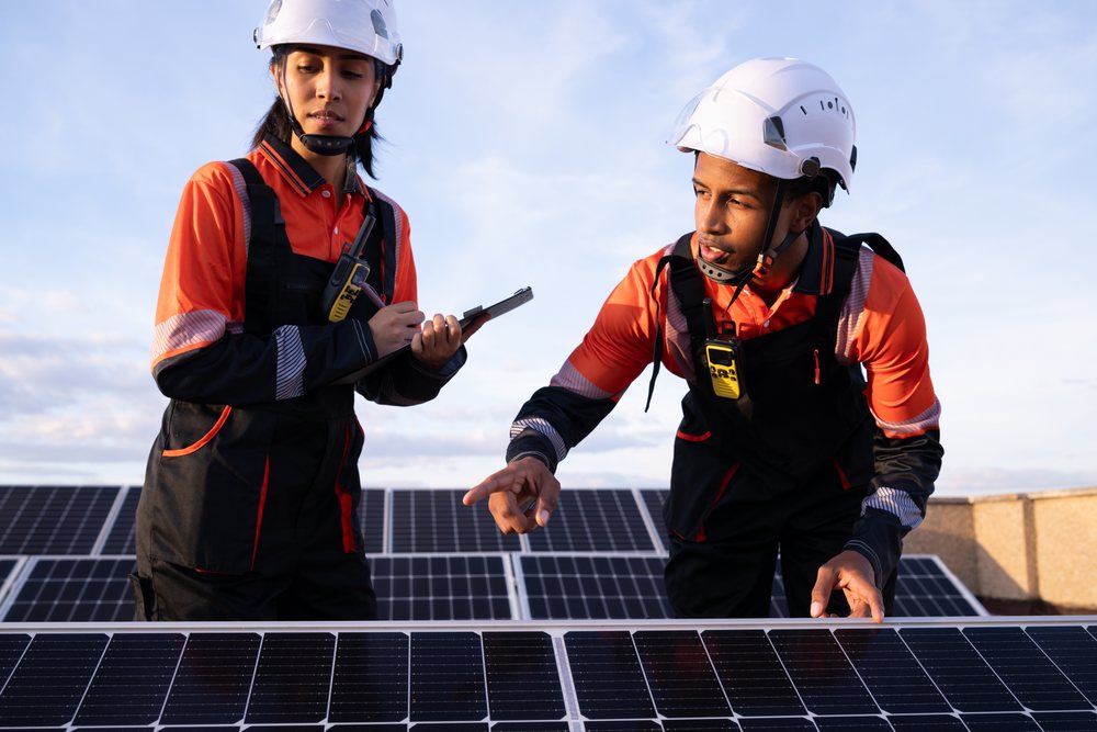 Two solar energy technicians inspect solar panels outdoors, taking notes and using radios, Madrid, Spain.