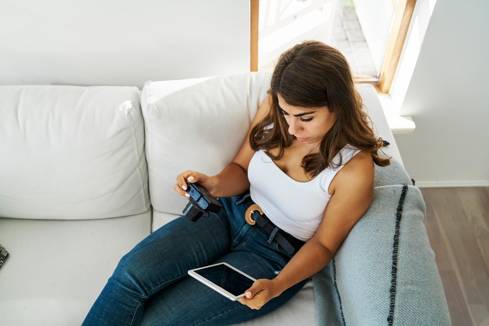 A white woman uses a digital camera and tablet while seated on a sofa