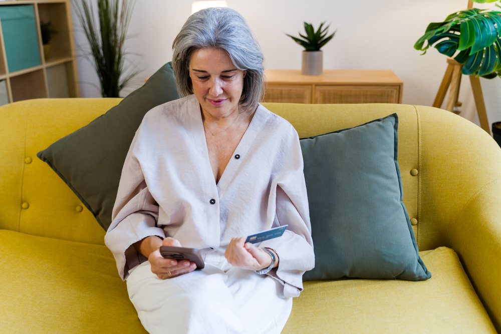 Mature woman with gray hair sits on a yellow sofa holding a smartphone and credit card in a modern living room, Spain.
