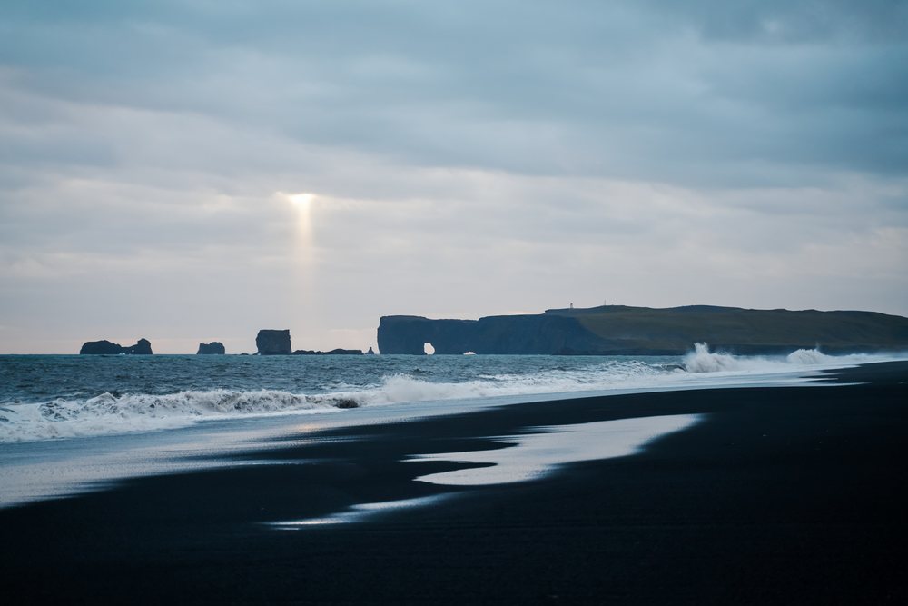 Waves crashing on a black sand beach with sea stacks under cloudy sky